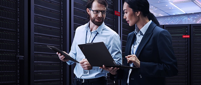 Two professionals reviewing information on laptops while standing in a data center aisle, with rows of rack servers in the background.