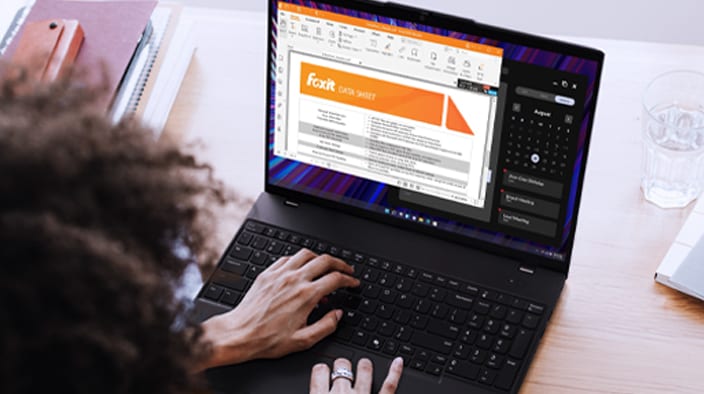 The image shows a person using a Lenovo laptop in a work environment. The laptop is open, and the screen displays a document editing interface, specifically a Foxit file, which includes graphics and text highlighted in orange. The person's hand is on the keyboard, suggesting they are interacting with the content on the screen.