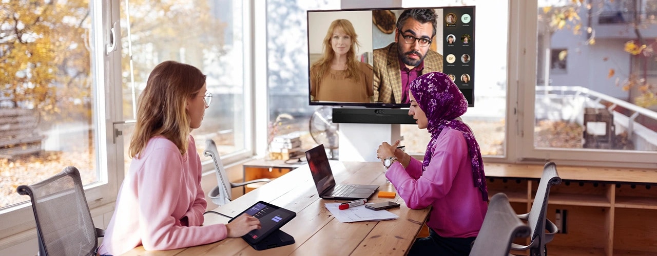 Teams call setting. Two in-room attendees: one using a Lenovo IP Controller on desk, the other looking at a Lenovo ThinkPad laptop, with ThinkSmart One for Microsoft Teams Rooms mounted on wall under monitor showing online attendees.