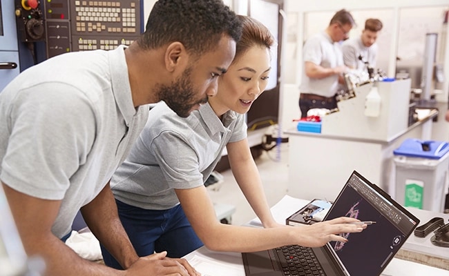 Two persons working on some graphics app opened on the Lenovo ThinkPad P16s Gen 3 (16 inch Intel) black touchscreen laptop placed on a desk in a technical lab setting with some other people working in blur background.