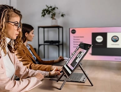 A person controlling a PPT via Lenovo ThinkBook Plus Gen 5 Hybrid (14” Intel) Tablet place on the easel stand & presenting it on a big screen monitor connected to the Hybrid Station, focusing productivity increase.