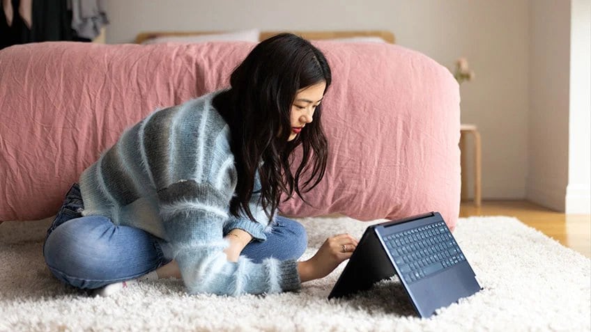 A woman sitting on a plush rug on her bedroom floor using a Yoga laptop