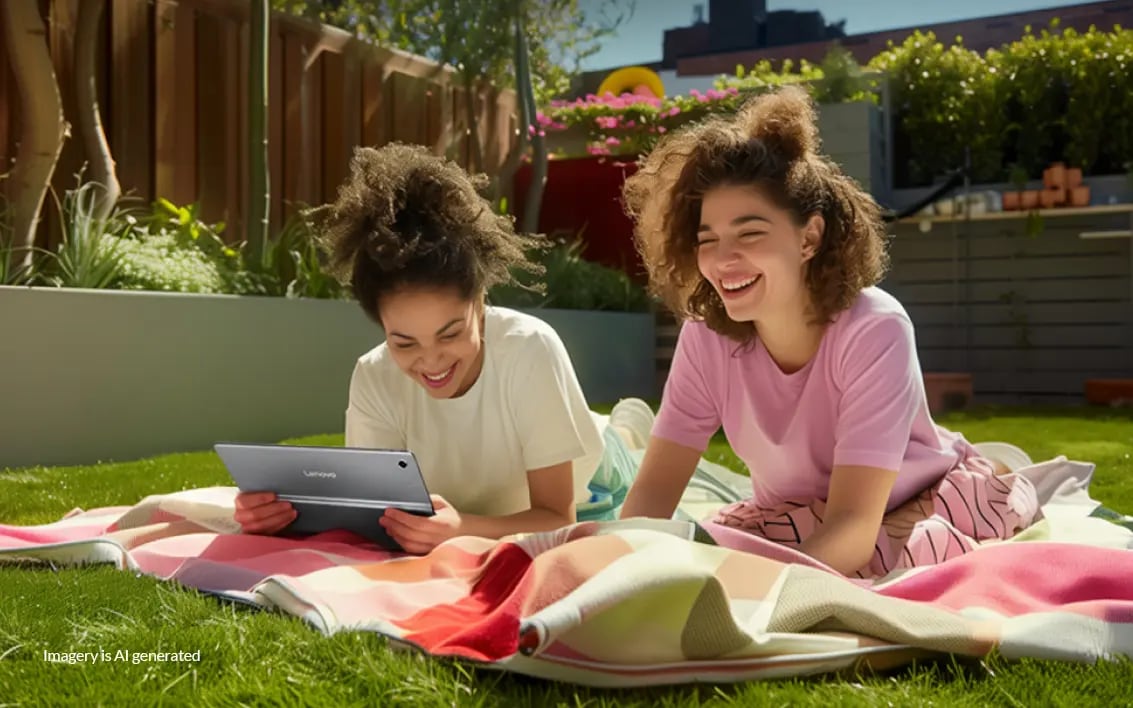 Two young women sitting on a blanket on top of grass, smiling and enjoying some time on a Lenovo tablet.