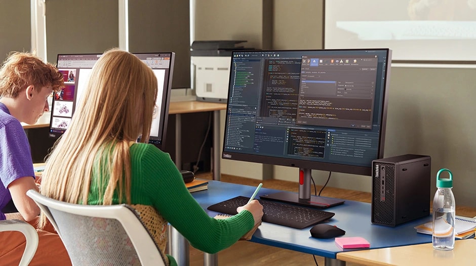 Two students at a desk, one taking notes as she looks at what appears to be code on a monitor connected to a Lenovo ThinkStation P3 Ultra SFF Gen 2 workstation