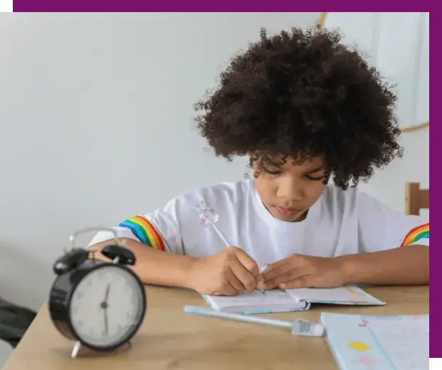 A child seated at a desk writes in a notebook. An alarm clock rests in the foreground.