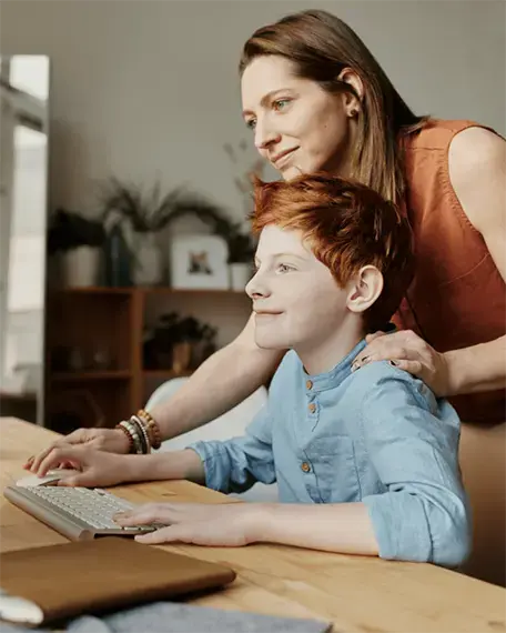 A woman rests her hand on a child’s shoulder as they both smile while looking at a computer display.