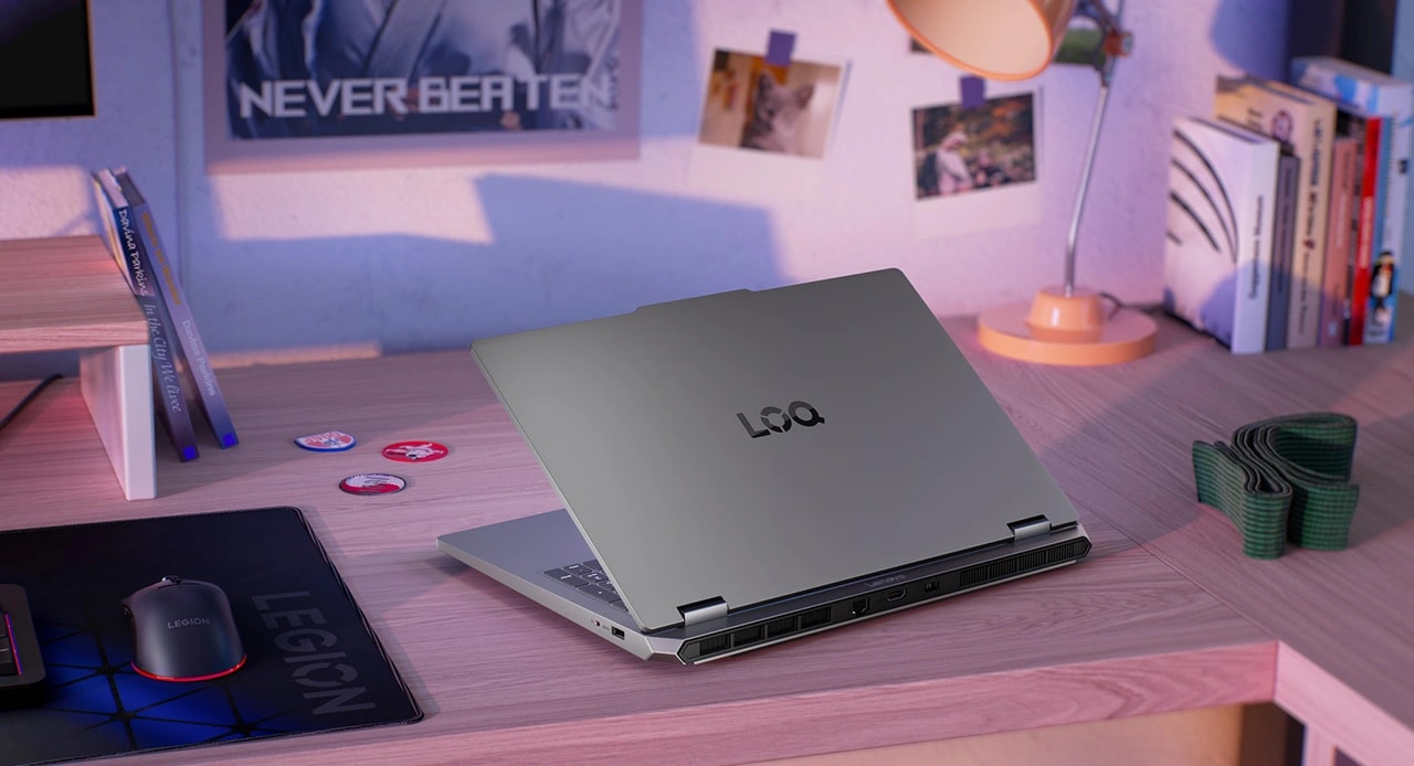 Lenovo LOQ Essential 15IRX11 gaming laptop on a desk with mouse, keyboard, mousepad, and monitor – rear view, lid partially open.