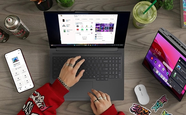 Top view of two hands typing on a laptop on a desk, surrounded by a phone, tablet, and mouse. The laptops is the Lenovo IdeaPad Slim 5a Gen 11 (16” AMD). מחשב נייד IdeaPad Slim 5 16AHP11 83S40013IV מבית Lenovo