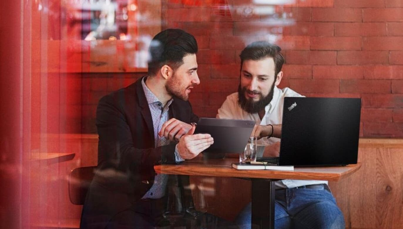 Two men in a casual workspace discuss content on a tablet, with a laptop on the table beside them.