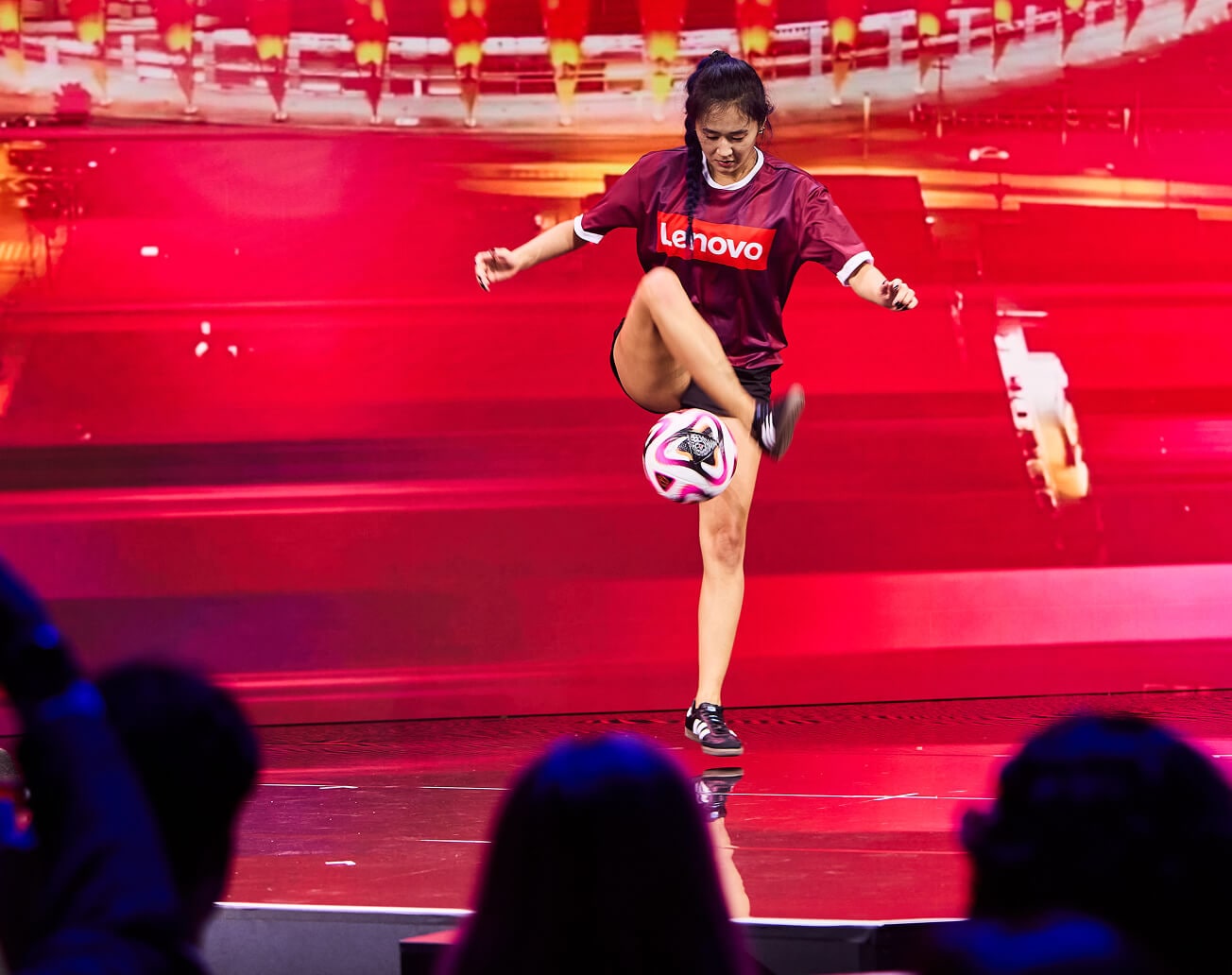 A performer in a Lenovo-branded shirt showcases soccer tricks on stage under bright red lighting, while attendees watch from the foreground.