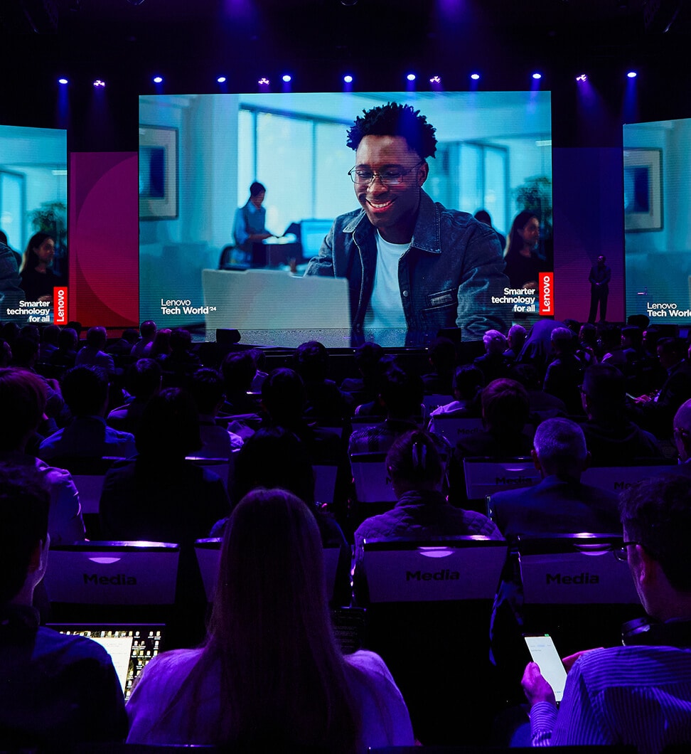 A large audience seated in a dark auditorium watches a Lenovo Tech World presentation on a big screen, featuring a person smiling at a laptop with the slogan 'Smarter technology for all.