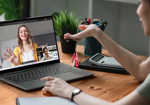 Two women speak on a video conference call, with one shown on the laptop screen while the other gestures with her hands.