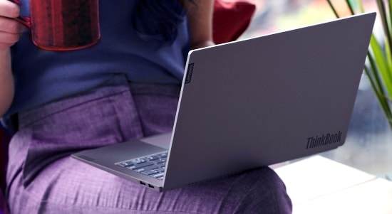 Une personne s’assoit avec un portable ThinkBook de Lenovo sur ses genoux, vêtus d’une jupe pourpre et d’un haut bleu, tenant une tasse texturée rouge dans une main. La scène est bien éclairée par la lumière naturelle et le couvercle du portable est partiellement ouvert, ce qui présente son design élégant gris argent et sa marque subtile.