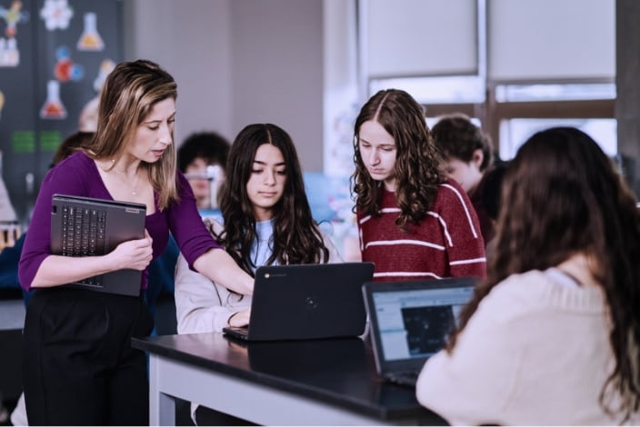 The image shows a classroom or lab setting with people working together on chromebook laptops, likely in an educational or instructional context.