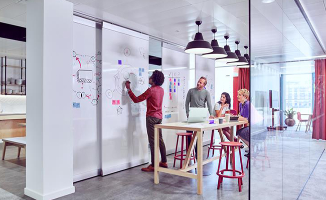 Team collaborating at a whiteboard in a modern office environment with laptop & tab placed on the adjacent office table.