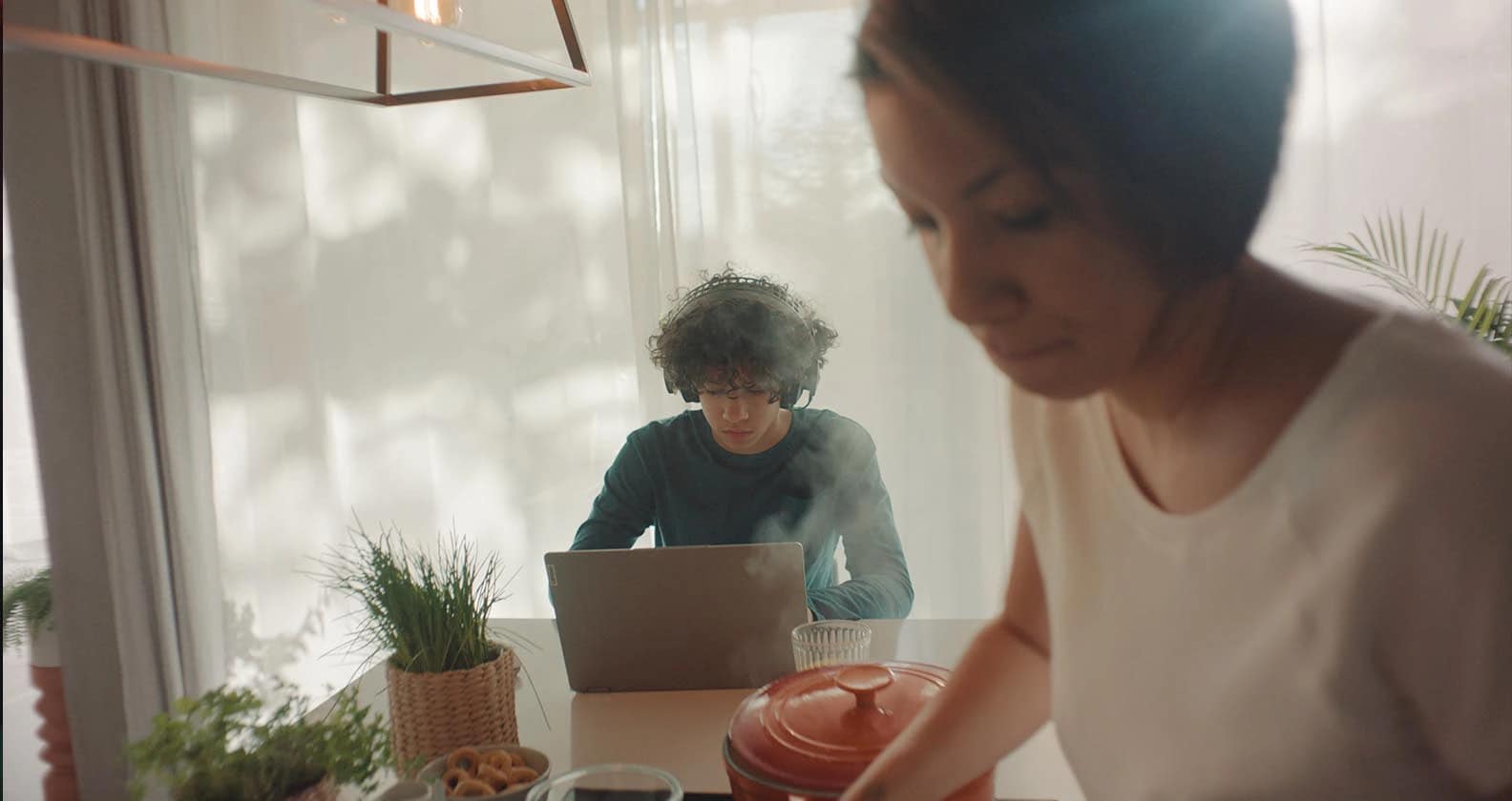 A teenage boy using his laptop on the kitchen table as his mother cooks in the foreground