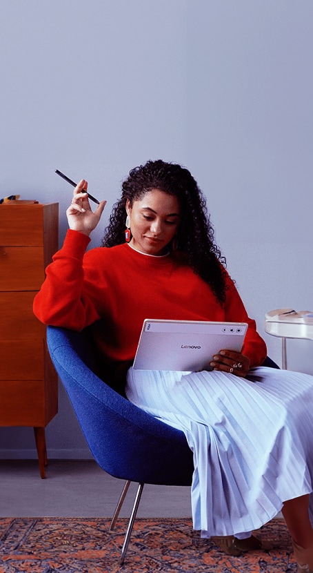 A woman sits on a blue chair holding Lenovo Tab Pen Plus and a Lenovo Yoga tablet, reviewing her art created on tablet.