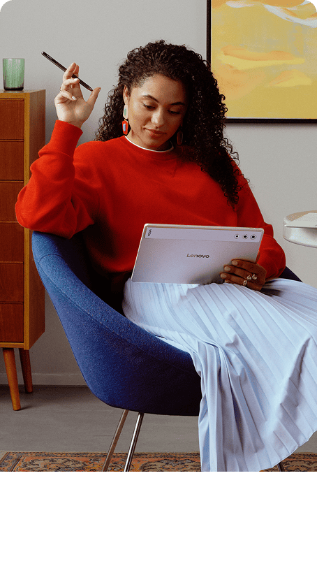 A woman sits on a blue chair holding Lenovo Tab Pen Plus and a Lenovo Yoga tablet, reviewing her art created on tablet.