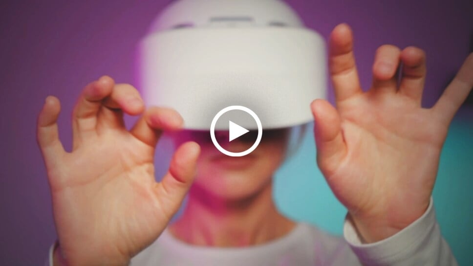 Picture of a young woman wearing a 3D Virtual Reality headgear. There's a play icon in the center of the image.