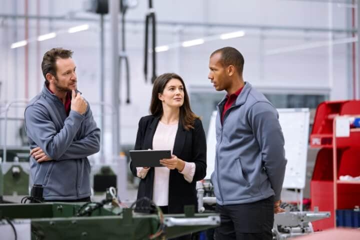 Three people are talking to each other while standing on the floor of a manufacturing plant.