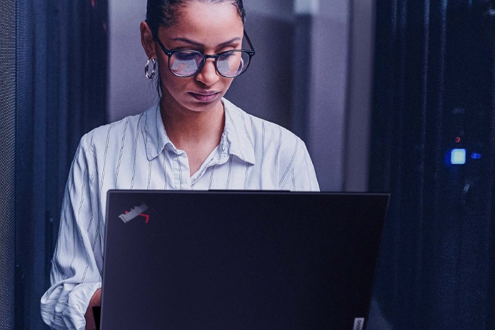 Image shows an IT tech standing inside a server room while looking down at a laptop.