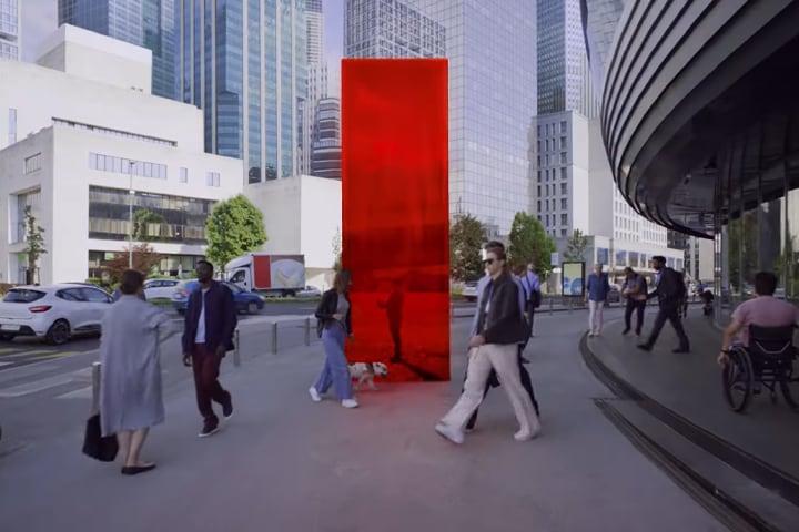 Image shows a busy street scene with pedestrians walking along a sidewalk where a vertical red slab is erected.