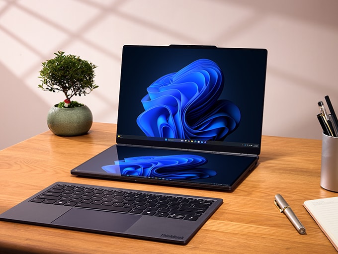 Atop a desk with a plant and various office supplies sits the Lenovo ThinkBook Modular AI PC concept with two pin-connected displays, both showing the blue Windows Bloom, and the detached keyboard sitting in front.