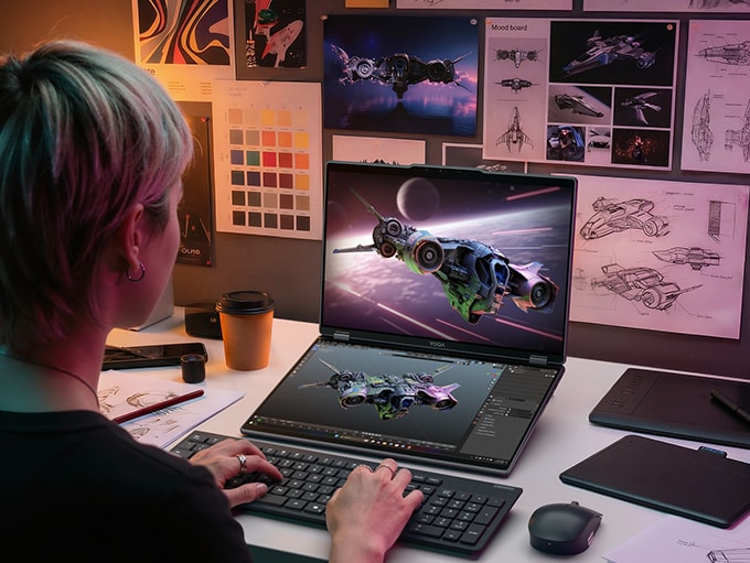 A person sits at a desk, using a wireless keyboard to work on a Lenovo Yoga Book Pro 3D 16″ laptop concept, which shows an illustrated spaceship on both displays.