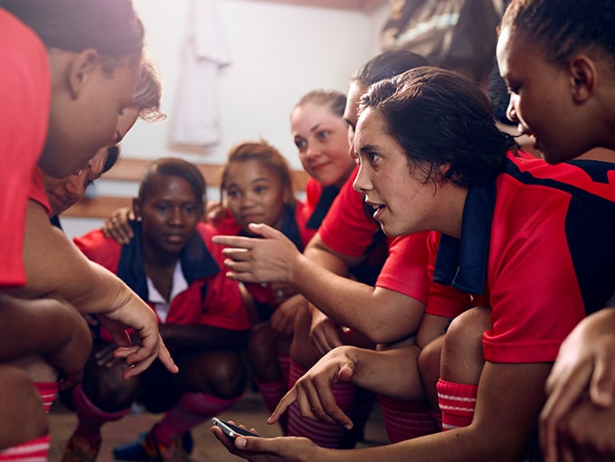 Members of a women's or teens' football/soccer team squat and lean into a huddle in their locker room, discussing the screen content of a mobile device.