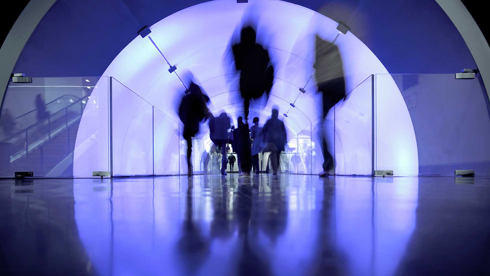 A group of people blurred out walking through a tunnel with transparent glass windows.