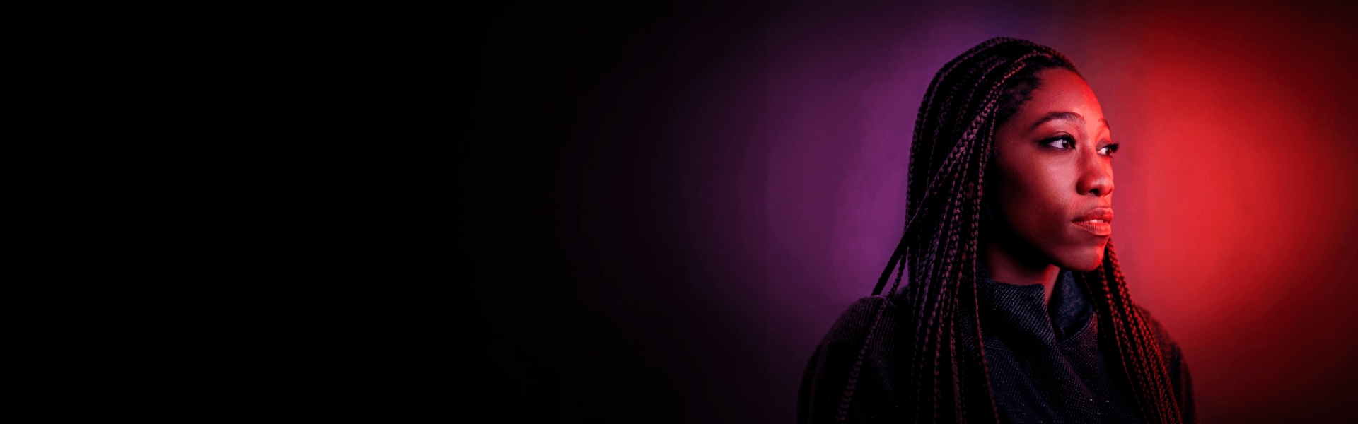 Woman with long braids against a dark background, lit by red light on one side.