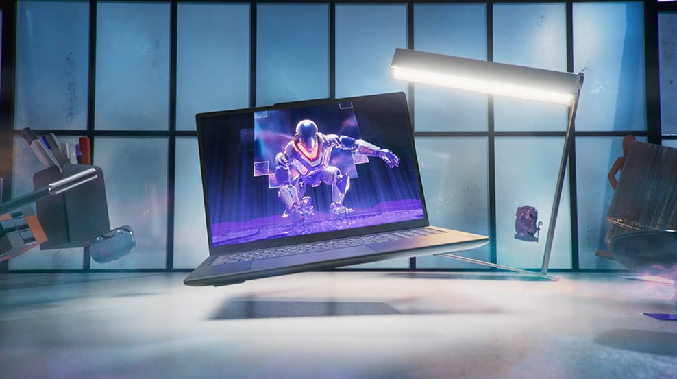 Un sac, une lampe, des fournitures de bureau et un ordinateur portable Yoga affichant l'illustration d'un personnage ou d'un robot futuriste planent au-dessus d'un bureau.