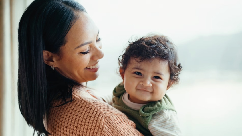 Mother holds her smiling baby in soft natural light.