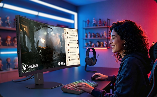 Woman smiles while gaming on a Legion 34w-2c monitor displaying Xbox Game Pass, with a headset on a stand and colorful figurines on shelves in a neon-lit room.