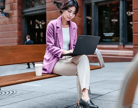 一位女士坐在木製長椅上，使用 Lenovo ThinkPad E14 Gen 7 (14 吋 Intel) 筆記型電腦，旁邊放著一個咖啡杯